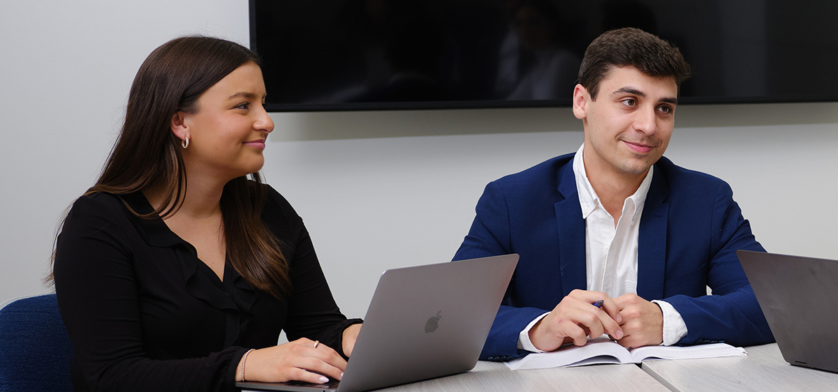 Two students sitting at a table, studying, one at a laptop, one with a textbook, both looking off-camera.